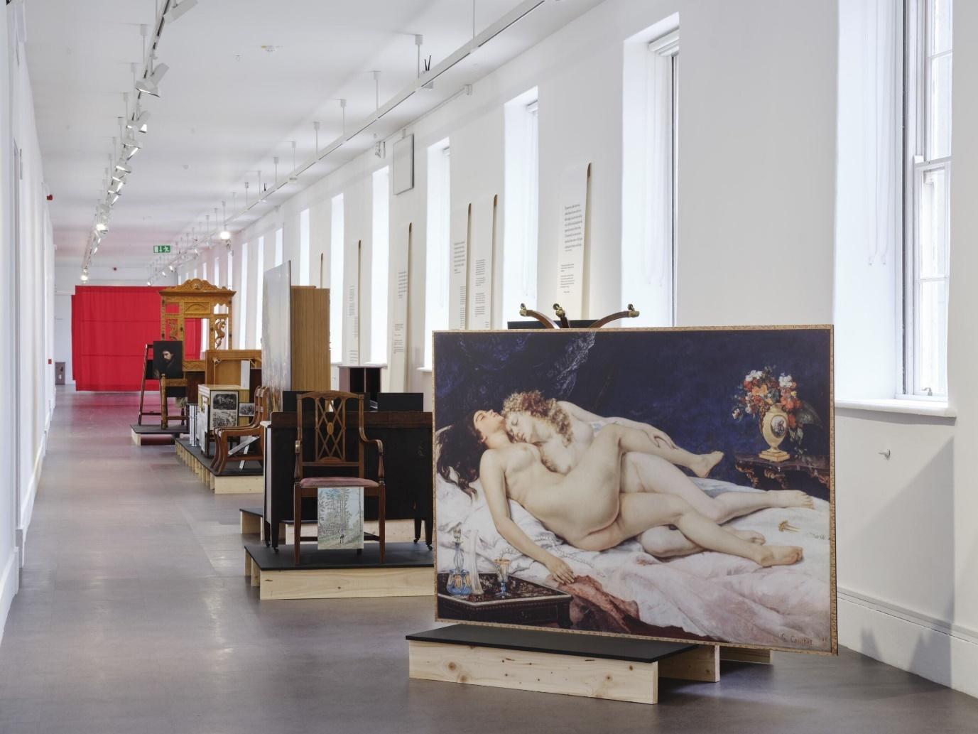 A long, well-lit gallery hall showcasing various artworks and antique furniture, including a large classical painting of two reclining nude figures in the foreground. The hall features wooden pieces and paintings displayed on platforms.