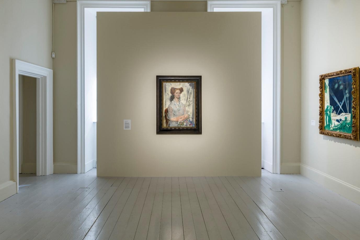 A single framed portrait painting displayed on a soft beige wall in a gallery. The painting shows a woman seated and holding a floral arrangement. Other colourful paintings hang on adjacent walls.