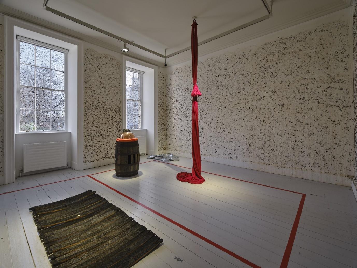 Art installation featuring a red aerial silk hanging from the ceiling, a barrel with an object on top, and abstract black and white wall patterns in a gallery room with wooden floors.