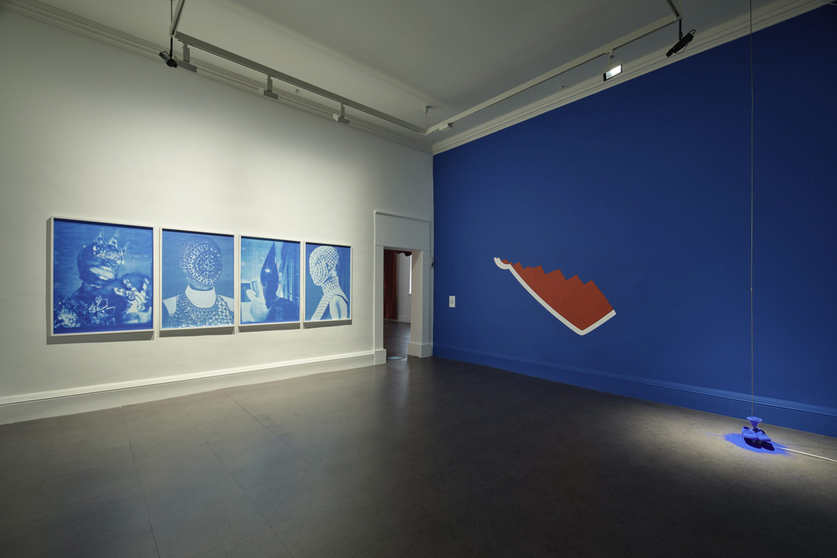 Interior installation photograph showing four blue framed prints on a white wall. The opposite wall is painted blue with a red mural that has been partially ripped off, the bottom of a red heart mural visable. On the ground is an installation of shoes and a funnel covered in blue pigment