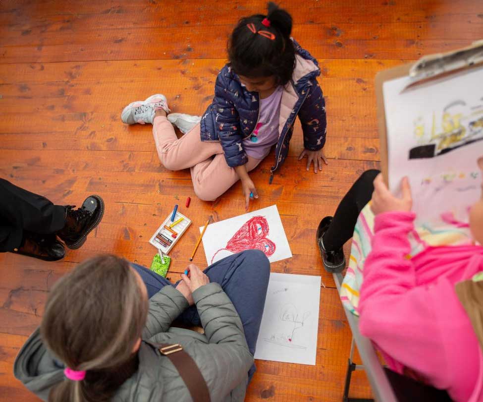 A young child sits on a wooden floor drawing with crayons on paper, while an adult, seated beside them, observes. The floor is scattered with children’s artwork, and another person holds a clipboard in the background.