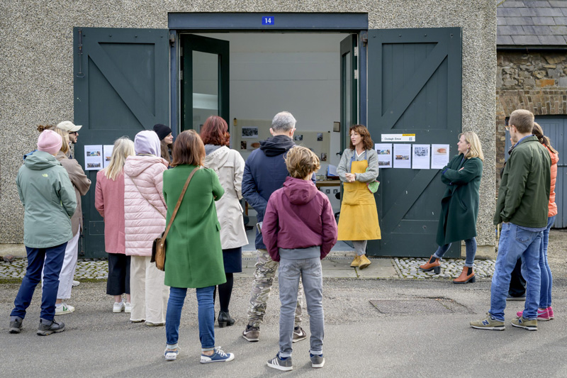 A photograph of fourteen people standing with their backs to the camera facing a white woman dressed in grey and wearing a long yellow artist’s apron who is addressing them, her hands clasped and her expression focused. She is standing in front of what looks like her studio space, with a table and some books visible inside, and pictures hanging on the back wall. Two large, dark wooden doors are opened wide, and inner glass-paned doors are opened inwardly. On the right-hand door, under a sign reading ‘Clodagh Emoe’ with ‘a visible plot’ written in smaller letters above, four A4 information sheets are displayed in a row, their content indiscernible. The left-hand door also has a row of sheets; only two are visible.