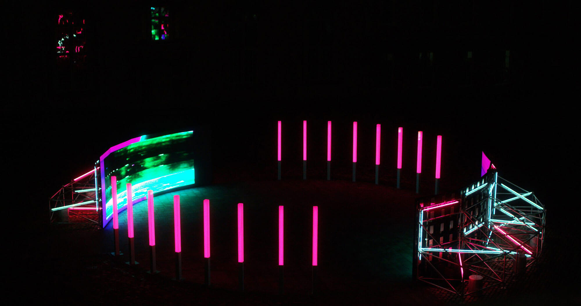 Night exterior installation photograph of a large screen displaying abstract neon colours. There are pink neon light columns on either side of the screen. In the foreground is the back of a second screen and it's supporting structure is illuminated.