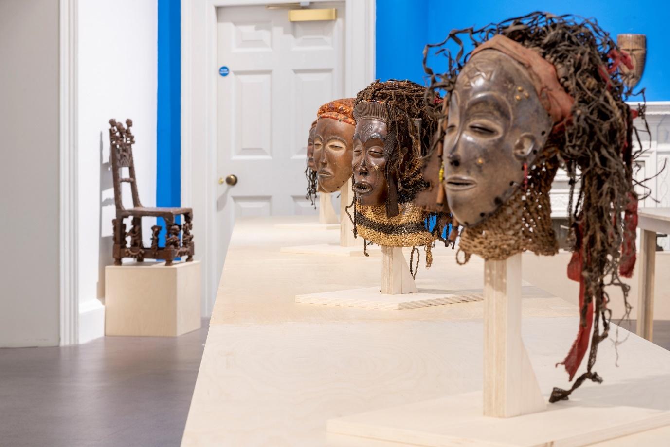 A gallery display of several intricately carved traditional masks mounted on stands, featuring natural fibres, beaded ornaments, and detailed facial features. In the background, there is an ornate wooden chair on a pedestal.