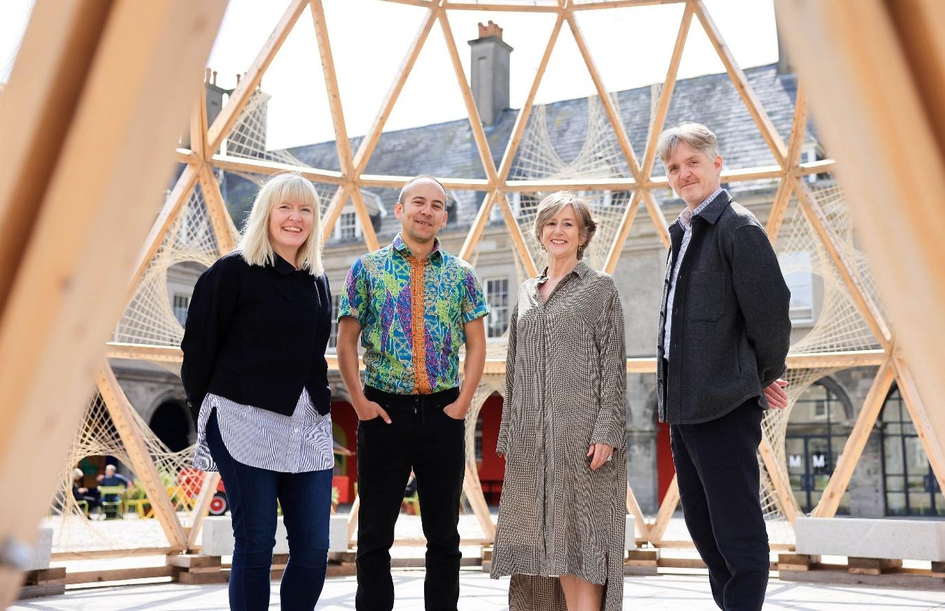 Four people stand outdoors in front of a large wooden geodesic structure. They are smiling, with two women on the left and two men on the right. The structure provides a geometrical backdrop, and the setting appears to be a public or cultural space.