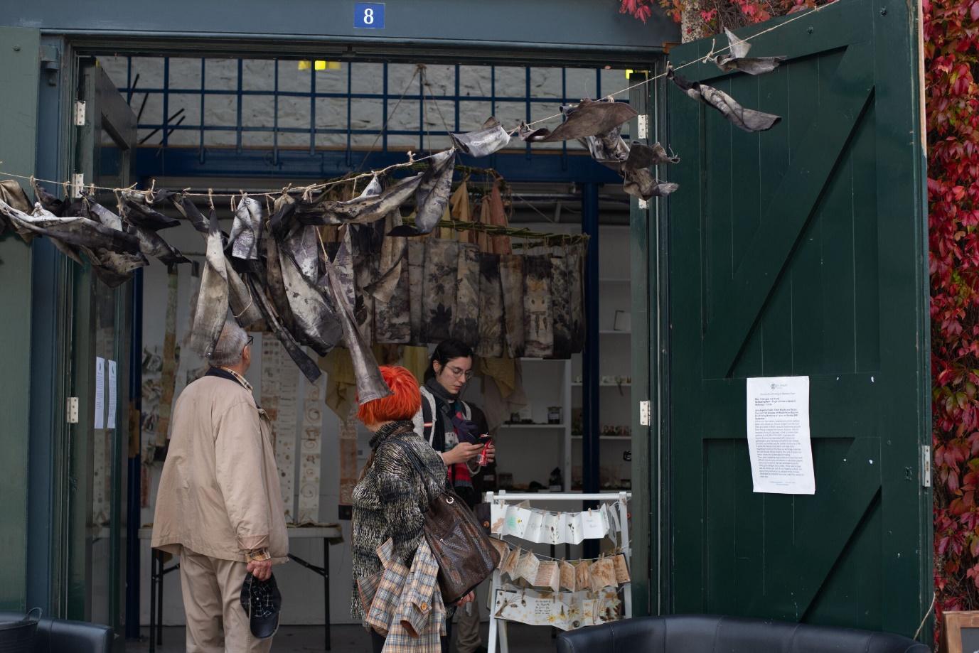 Three people stand outside the entrance of a workshop or gallery, looking at an art installation. Above the doorway, there is a string of fabric strips or leaves hanging, and the green wooden door is open, revealing more artworks