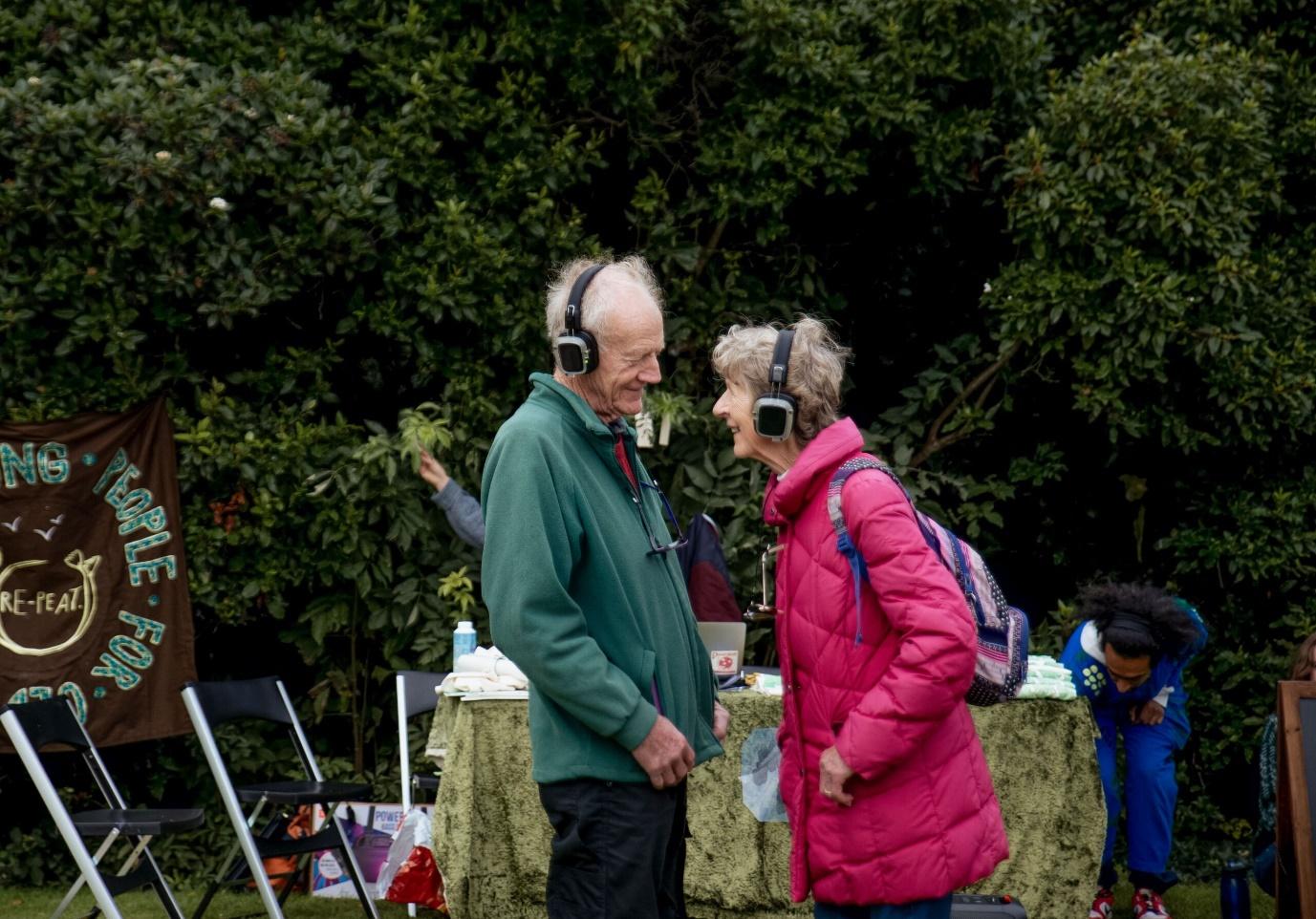 An elderly man and woman stand outside, smiling and interacting while wearing wireless headphones. Behind them is a table with a green cloth and a banner that promotes environmental awareness, along with chairs set up for what appears to be an outdoor event.