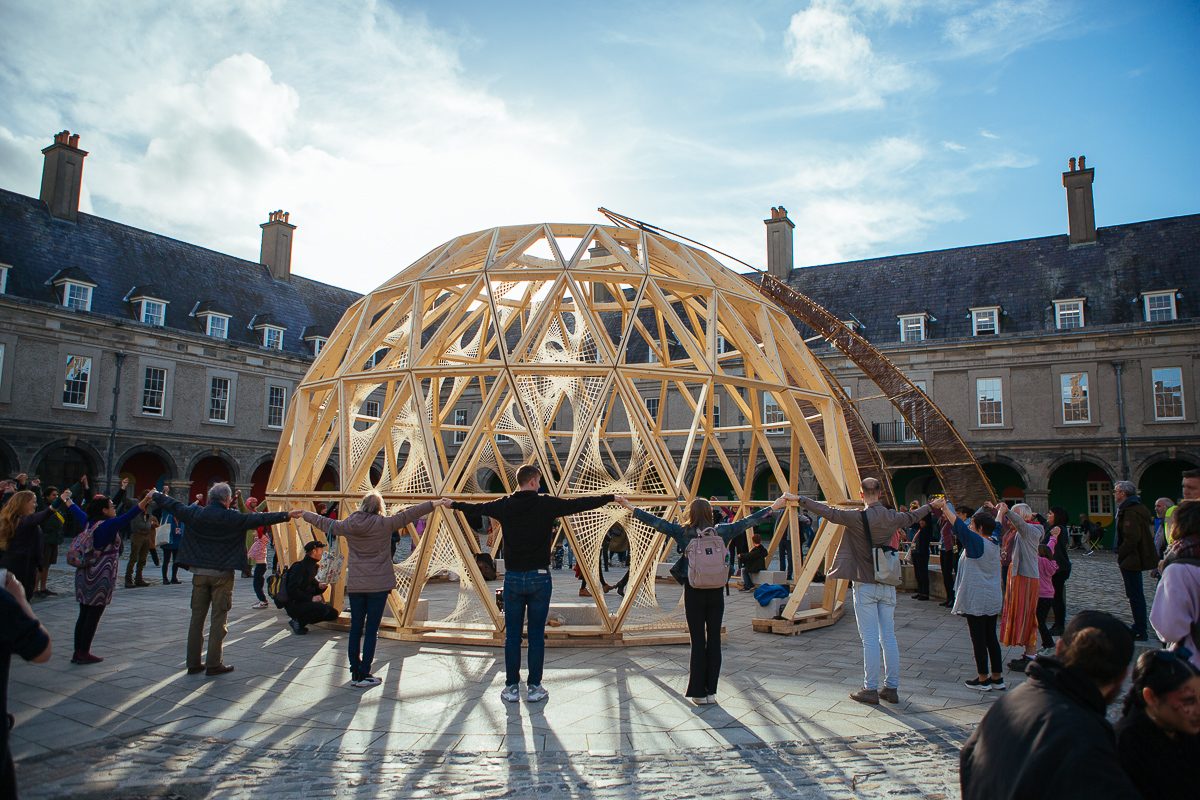 A group of people standing in a circle holding hands around a large wooden geodesic dome structure in an open courtyard. The sky is partly cloudy, and the surrounding buildings are historic with tall chimneys