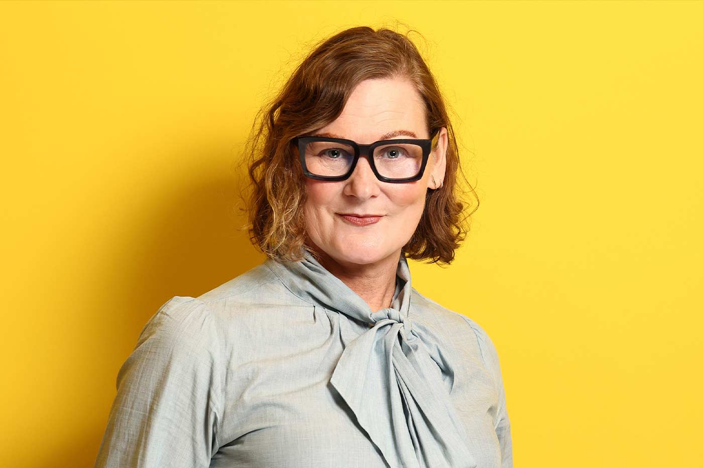 A portrait of a woman with shoulder-length curly hair wearing a light blue blouse with a bow and black trousers. She is smiling and standing in front of a bright yellow backdrop, her hands casually in her pockets.