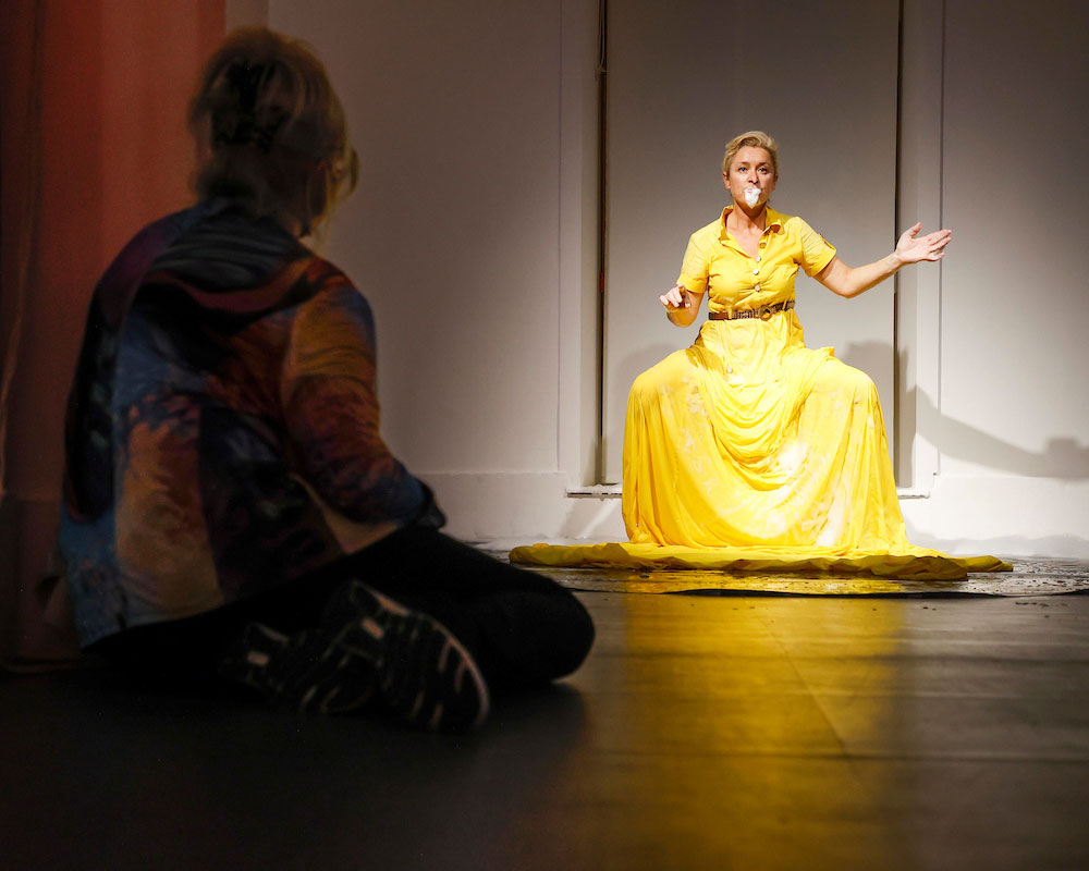In this photograph, a woman sits in shadow with her back to the camera on a polished floor watching the performance artist Amanda Coogan create her piece ‘Yellow’. Amanda, a spotlit white woman with blond hair wears a long yellow dress that trails the floor in front of her, and appears to be sitting on something we can’t see in a window or doorway. She appears in a trance, her knees wide apart, her left arm is slightly bent and out to the side. Her right arm, also bent, points casually towards the observer. The folds of the fabric in the skirt are accentuated and you realise that they are dotted with soap suds. Her mouth is stopped with what could be white fabric, or more soap suds could be bubbling out from within her.