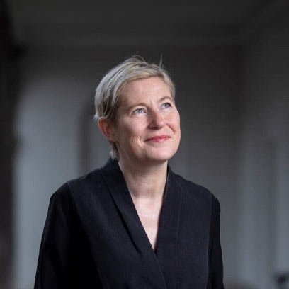 Annie Fletcher, Director of IMMA, A woman with short blonde hair, wearing a black outfit, looking upwards with a thoughtful expression, standing in a softly lit indoor space.