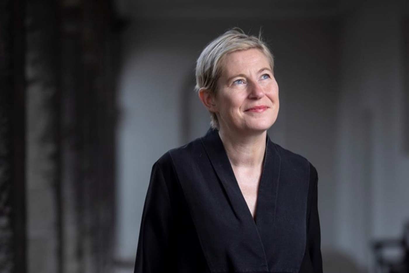 Annie Fletcher, Director of IMMA, A woman with short blonde hair, wearing a black outfit, looking upwards with a thoughtful expression, standing in a softly lit indoor space.