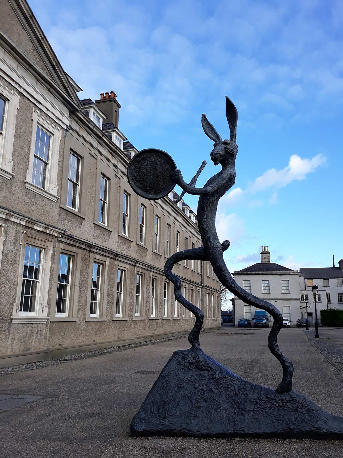 A sculpture of a tall, thin hare with elongated ears, standing on two legs and holding a circular object in one hand. The sculpture is outdoors, with a historic building and a blue sky in the background.