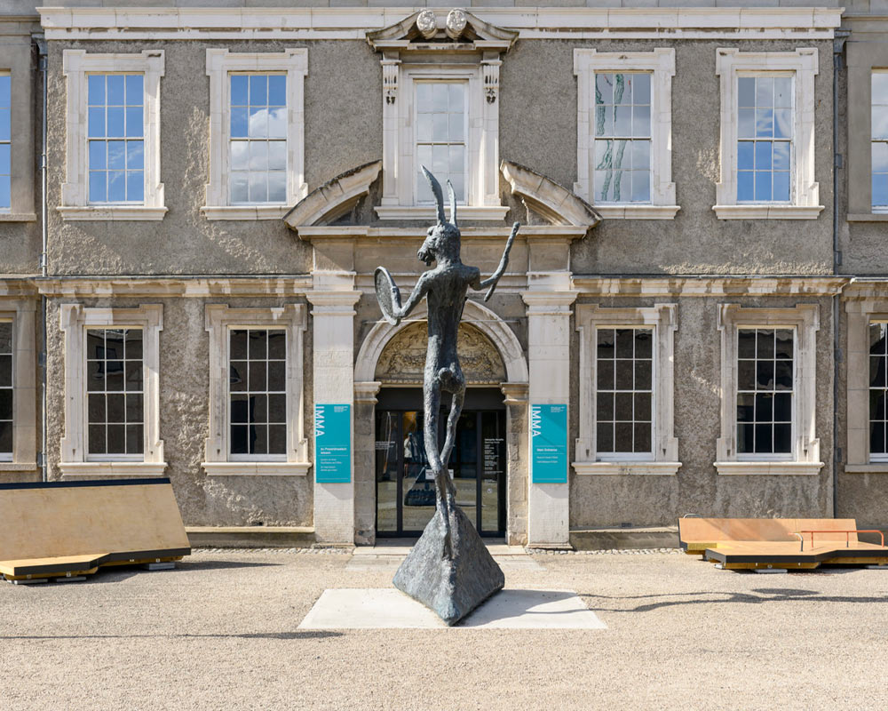 A photographed view of the front entrance of IMMA, on the South side of the Royal Hospital Kilmainham, with blue sky reflected in the top 7 windows. The facade is 17th century Classical, apart from the modern, glass door, in which the reflection of the photographer is just visible. The three ground floor windows to either side of the front door are almost in darkness. ‘The Drummer’, a grey bronze sculpture by Barry Flanagan, of a tall hare standing on a triangular base is holding a stick in one hand/paw and a round, drum-like object in the other, as though about to beat the drum. The hare’s ears reach as high as the upper floor of the building. To the left is a structure that resembles a wooden door, propped length-ways on its side, creating a triangle shape on a base. To the left of that is a grey, square planter holding a green conical-shaped box-hedge. To the right of the hare is another wooden structure that looks like low seating behind a low platform with a small, red metal railing at the front, and there is an identical planter with conical-shaped box-hedge to the right of that. The two seating structures are installations created by the artists’ collective Forerunner. The hare casts a long shadow to the right across the gravelled forecourt.