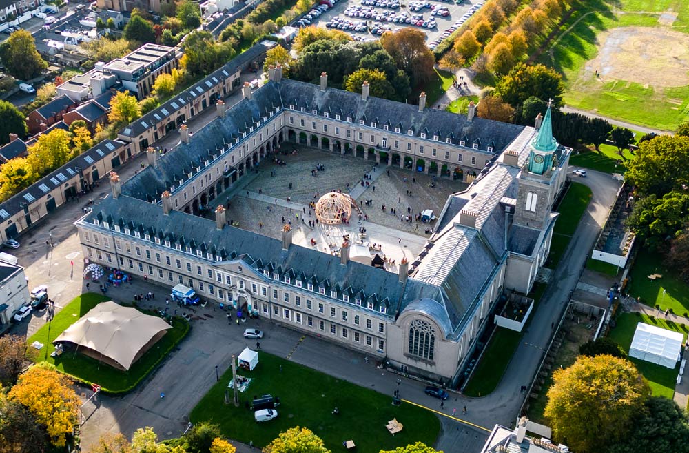 A photographed aerial view of IMMA with its inner colonnaded courtyard clearly visible. The courtyard is divided into quadrants by four paths that meet in the centre. Right in the middle is what is described as an eco-pavilion, which was part of the Earth Rising Eco Art Festival in 2022. It is a spherical structure made of a willow lattice through which people can be seen wandering about. To the right is a flap constructed of the same material, attached to the top of the structure, and raised off the ground on solid blocks. People are strolling about the courtyard, and to the right, in front of the clock tower, there is a van-like structure that could be a food outlet. People can be seen sitting at wooden picnic tables. The East wing of the building is in the foreground, with the Baroque Chapel stained glass window to the right. The Classical-style building includes a clocktower surmounted by a blue-green pointed copper pinnacle, with a weathercock. There is a beige bulging tent facing the facade in the foreground, with other tents and a few vehicles dotted about. Rows of houses, a car park and a sports field can be seen in the background.