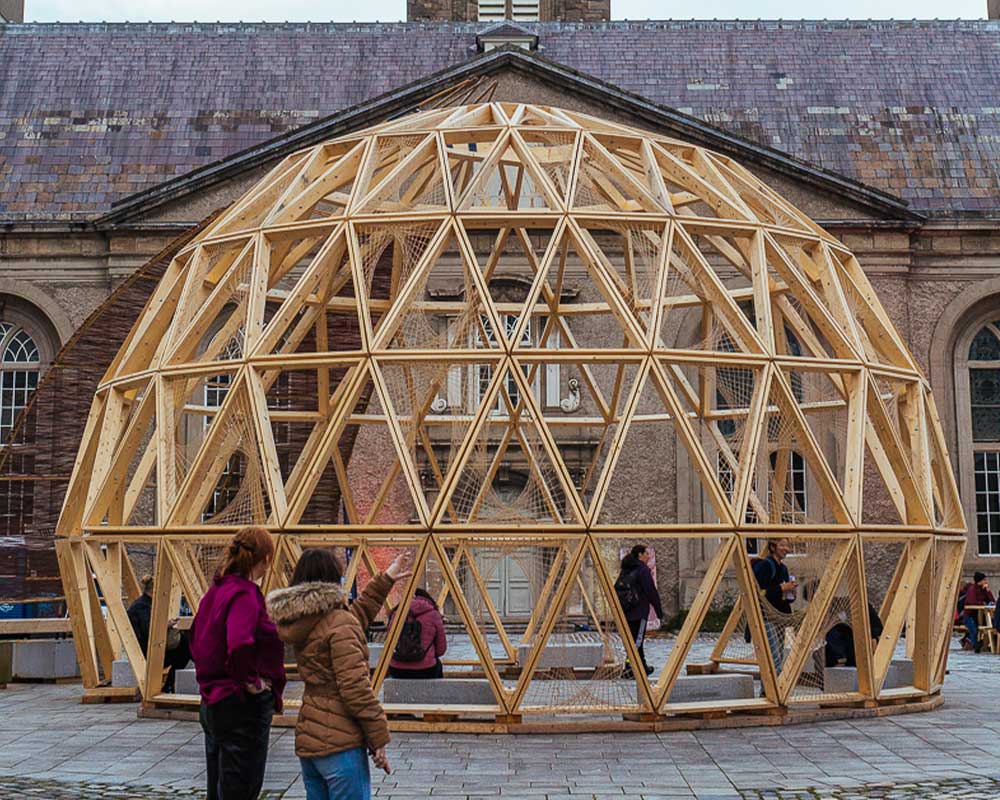 A photographed view of the courtyard at IMMA with the North Range partially obscured by a temporary installation, the Eco-Pavilion, a large, almost spherical lattice construction made of willow. Two people wearing winter jackets stand in the foreground with their backs to us discussing the piece. Three people can be seen inside the structure, which is perfectly centred under IMMA’s green-blue clock tower, with its sharply pointed copper spire rising into a slightly hazy blue sky behind.