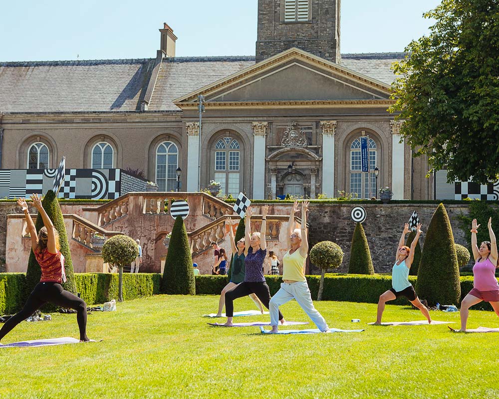 This is a photograph of a yoga or exercise class taking place on a sunny day in the Formal Garden at IMMA. Five white women reaching towards the sky, lunging with their front legs, face their teacher, also a white woman. They stand on yoga mats in warrior position. The lawn they are on is surrounded by a low box-hedge with alternating conical bushes and round topiary bushes just inside it. Some of the bushes have black and white geometric shapes on top of them. In the background, the classical double stone staircase leads to one of IMMA’s terraces, part of which is obscured on the left by a hoarding covered in similar black and white geometric patterns. We can see the base of the clock tower rising into a hazy blue sky.