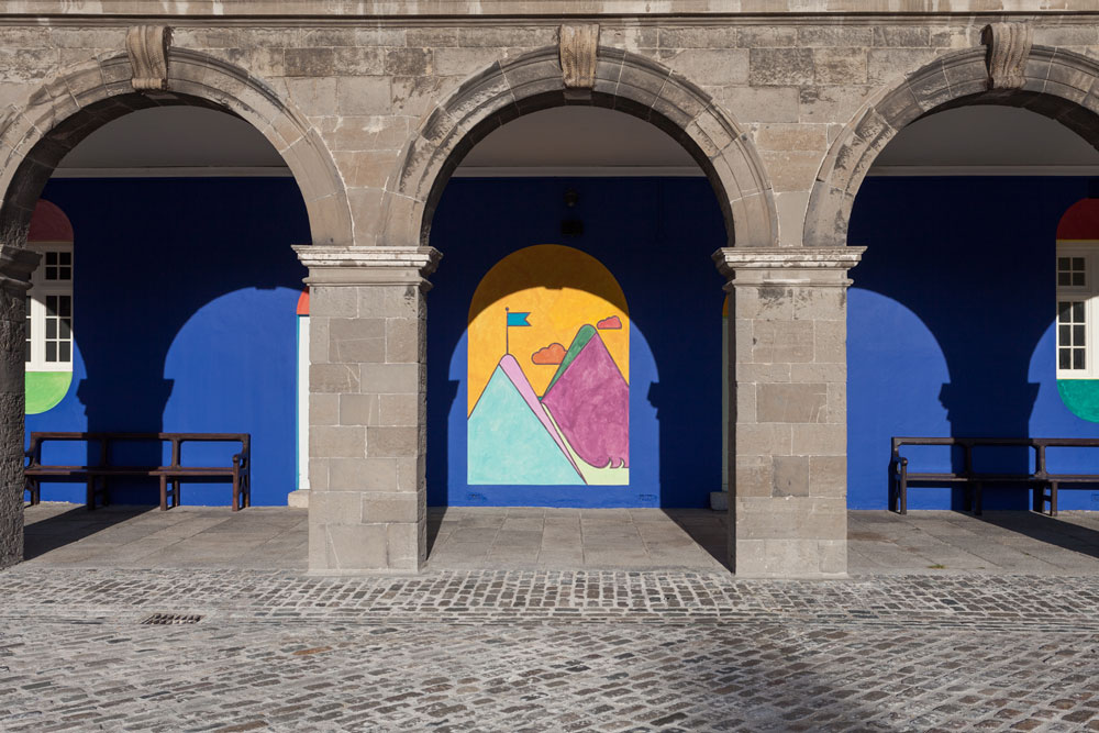 A photographed section of one side of the IMMA’s courtyard, taken at an angle so that the right-hand side of the image is taken up by one arch of which only the left supporting pillar can be seen, and four diminishing arches are visible to the left. Through the arches, site specific paintings by Navine G. Dossos are visible on the colonnade wall. The installation is called ‘Kind Words Can Never Die’, and this view features a red background. Through the closest arch there is a white door with a stone doorstep and a pale blue semicircle painted over it, in a fanlight shape. There is an arched-shaped painting to the left of the door of two sets of three overlapping ovals, mirrored vertically to make a flower shape, with a circle drawn in the centre of each set. Each of the sections created by the overlaps is a different colour: yellow, green, red, and white on top, and peach, green, black, white, and two shades of blue on the bottom. The background is mottled dark blue. Through the second arch is a white window with a purple semi-circle above, and an orange semi-circle below. It’s edged at the side in green. Beneath the window are two narrow benches with arms and backs. The third arch reveals another, similar white door with a dark green fanlight painted above. The angle of the image means that the viewer cannot see through the remaining two arches.