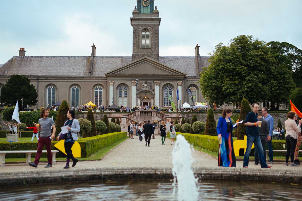 A wide view of a large building with a clock tower, surrounded by a beautifully manicured garden. People are walking along the path, and there are tents and booths set up on the terrace in front of the building, indicating a public event or festival.