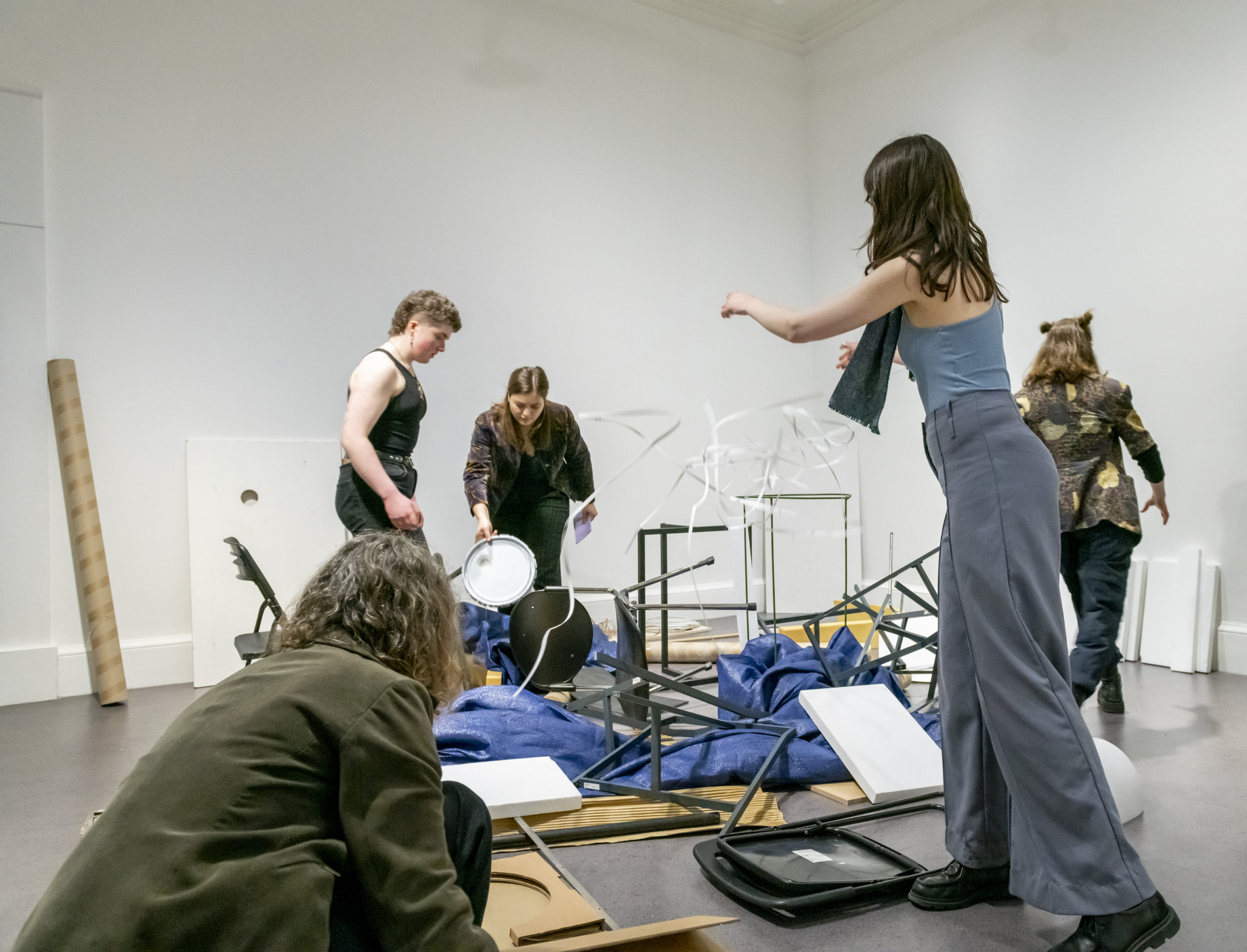 A group of people assembling or disassembling an art installation inside a gallery space, working with various materials scattered on the floor. They appear to be collaborating on the structure.