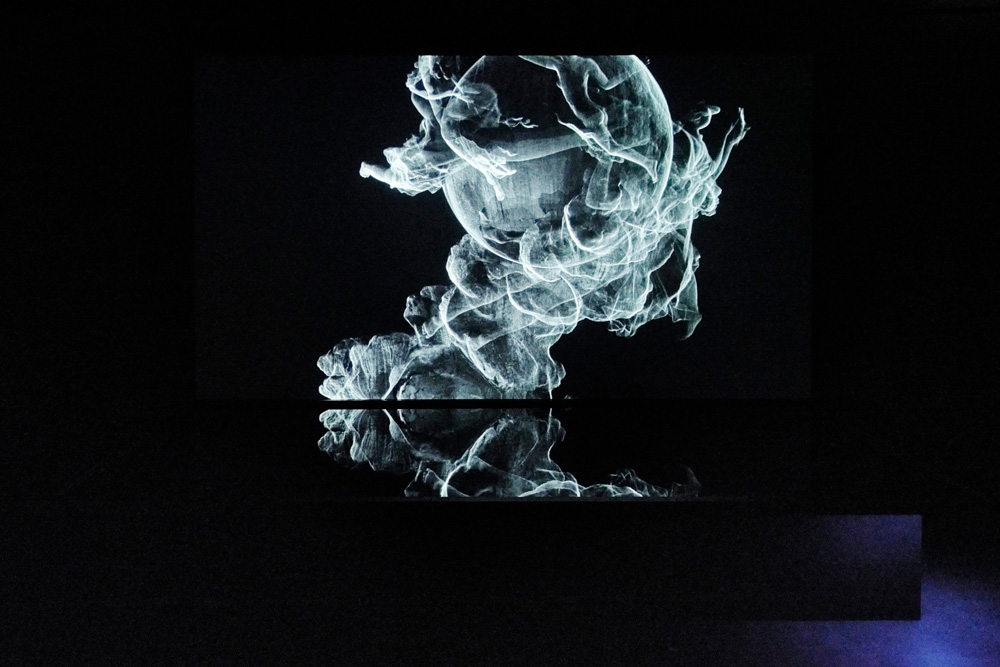 A computer animation projected in a dark room, looks at first like wisps of smoke against a black background. It is in fact black and white image of figures flying around a central globe. A black reflective plinth or perhaps a container of water below mirrors some wisps of the projected image. In the bottom right corner, a light we cannot see picks out the corner of the plinth and a patch of floor, which looks blueish, while everything else is black. The piece is ‘Sovereign Sisters’ by the Otolith Group artist collective’s exhibition Xenogenesis.