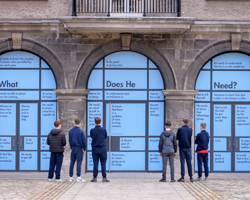 This photograph shows a pair of modern double doors at IMMA with pale blue panels divided by dark grey frames. Dark grey writing in the two upper panels says, on the left, “Women to show him how to be a man.” and on the right “Money, power and a bigger dog.” The door handles are vertical metal bars. To the right of the handle on the right-hand door the panel says “To be heard.” The panel to the left is obscured by a white man with dark hair who can be seen from the hips up, standing with his back to the camera. He is wearing a plaid shirt and is using a mobile phone to capture a QR code stuck just above the handle on the door to the left. The blue panels are part of an installation at IMMA called ‘What Does He Need?’.