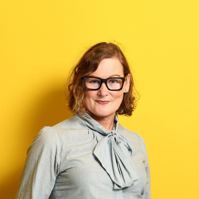 A portrait of a woman with shoulder-length curly hair wearing a light blue blouse with a bow and black trousers. She is smiling and standing in front of a bright yellow backdrop, her hands casually in her pockets.