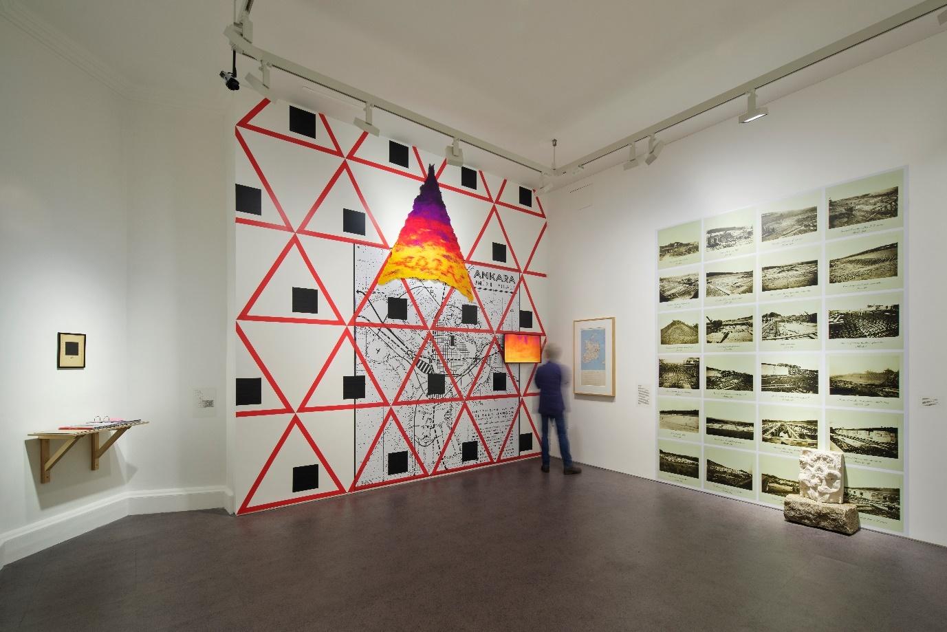 Art installation featuring a red and black geometric mural and a series of landscape photographs displayed in a gallery.