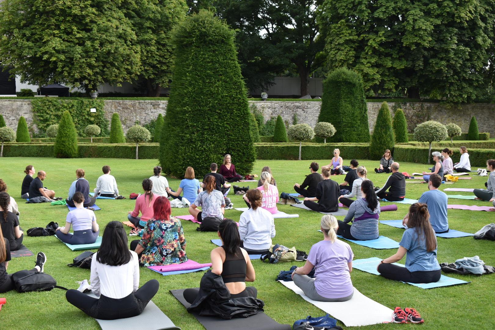 A group of people participating in an outdoor yoga session, sitting on mats in a well-manicured garden surrounded by neatly trimmed shrubs and trees. The instructor sits in front, leading the class.