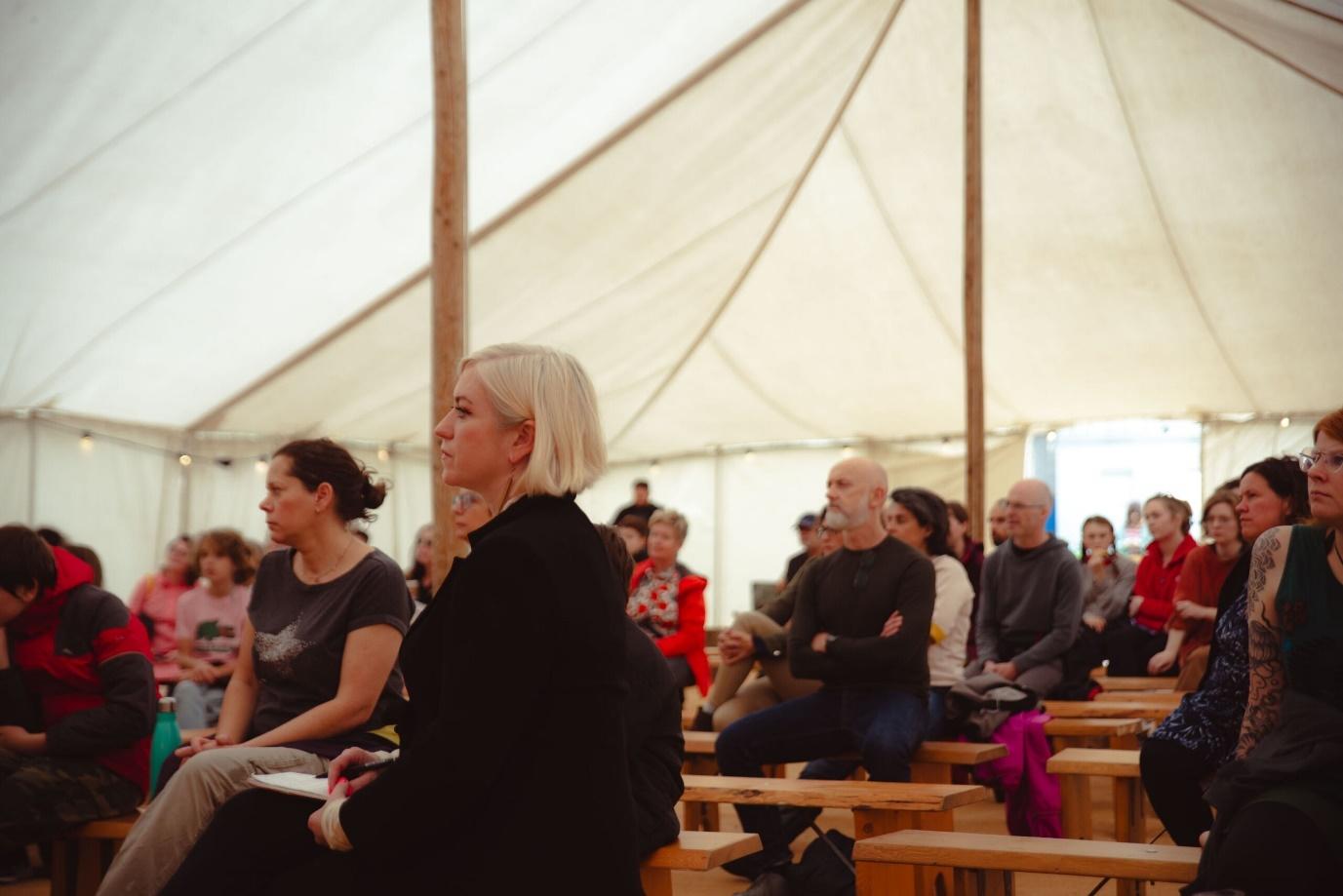 A group of people seated on wooden benches inside a large tent. They appear to be attentively listening to a presentation or talk, with a focus on a woman with short blonde hair seated near the front.