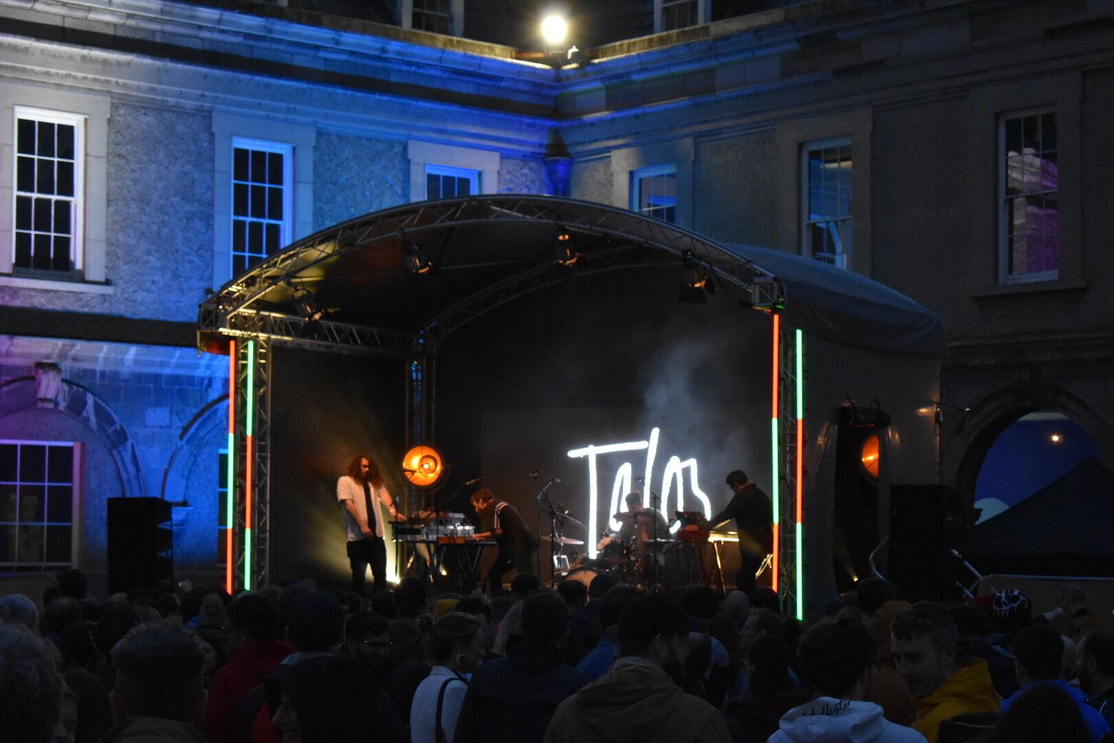 An evening courtyard concert at a historical building, illuminated by purple lights. A crowd is gathered in front of an outdoor stage, with a clock tower and cloudy sky visible in the background