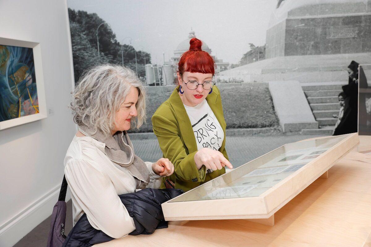 Two women standing inside a gallery, looking at a glass display case. One woman with grey hair listens as the other, with red hair and glasses, points at something inside the case.