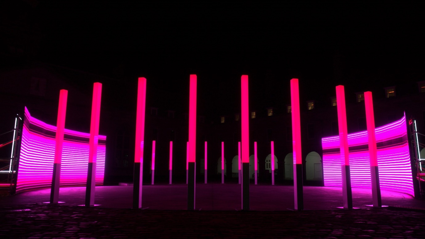 Night exterior installation photograph of two large, parallel, screens that are neon pink in colour. There are pink neon light columns on either side of the screens.