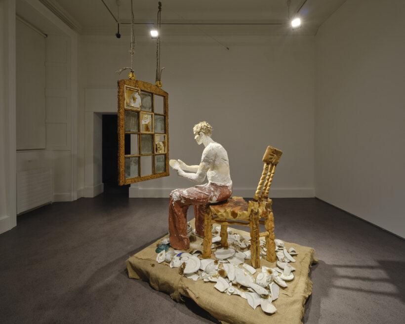 A sculpture installation of a seated figure made from a plaster-like material, surrounded by broken ceramic plates and bowls. Above the figure is a suspended, rustic window frame with glass panes.