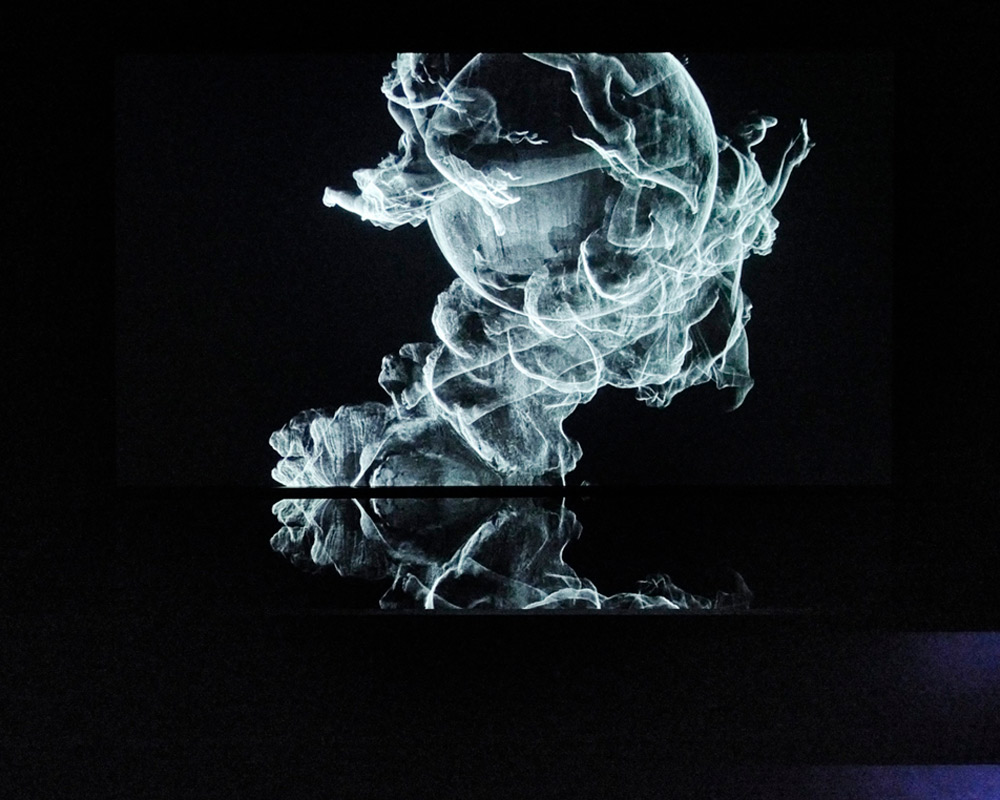A computer animation projected in a dark room, looks at first like wisps of smoke against a black background. It is in fact black and white image of figures flying around a central globe. A black reflective plinth or perhaps a container of water below mirrors some wisps of the projected image. In the bottom right corner, a light we cannot see picks out the corner of the plinth and a patch of floor, which looks blueish, while everything else is black. The piece is ‘Sovereign Sisters’ by the Otolith Group artist collective’s exhibition Xenogenesis.