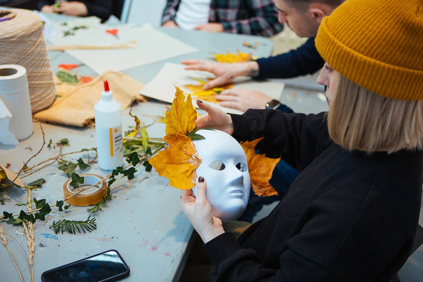 A person wearing a mustard yellow beanie is decorating a white mask with autumn leaves and twigs. Others are working on crafts in the background, with various craft materials scattered on the table, including glue, tape, and a large spool of twine