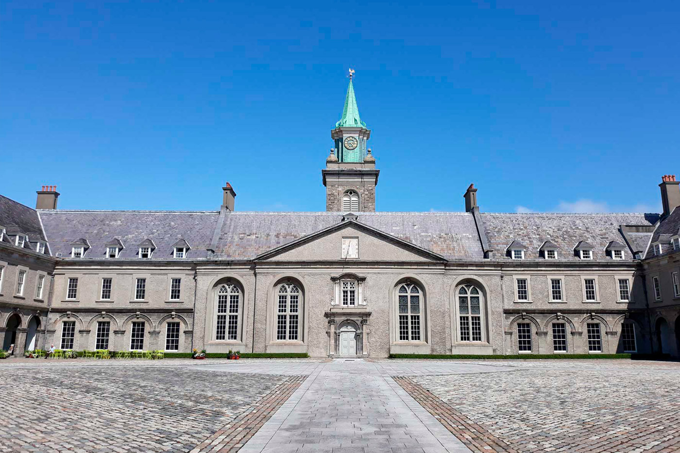 The image shows the facade of the Irish Museum of Modern Art (IMMA) in Dublin, Ireland. The building is symmetrical with a central section that has a prominent clock tower topped with a green spire. The structure is characterized by its gray stone exterior, large arched windows, and a cobblestone courtyard in the foreground.